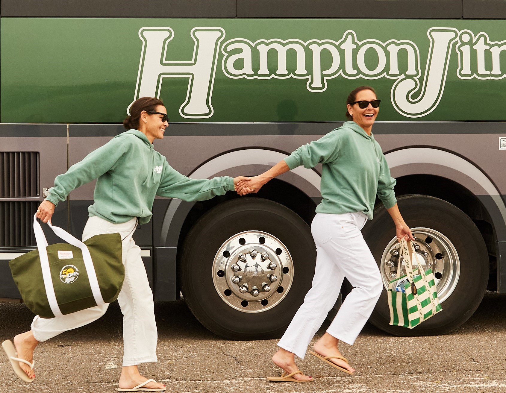 Two friends in matching green hoodies playfully pose while holding hands in front of a Hampton Jitney bus.